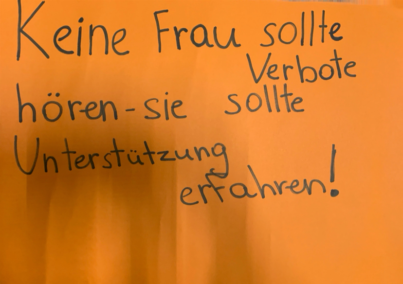 Orangefarbenes Schild mit Text: Unterstützung statt Verbote für Frauen.